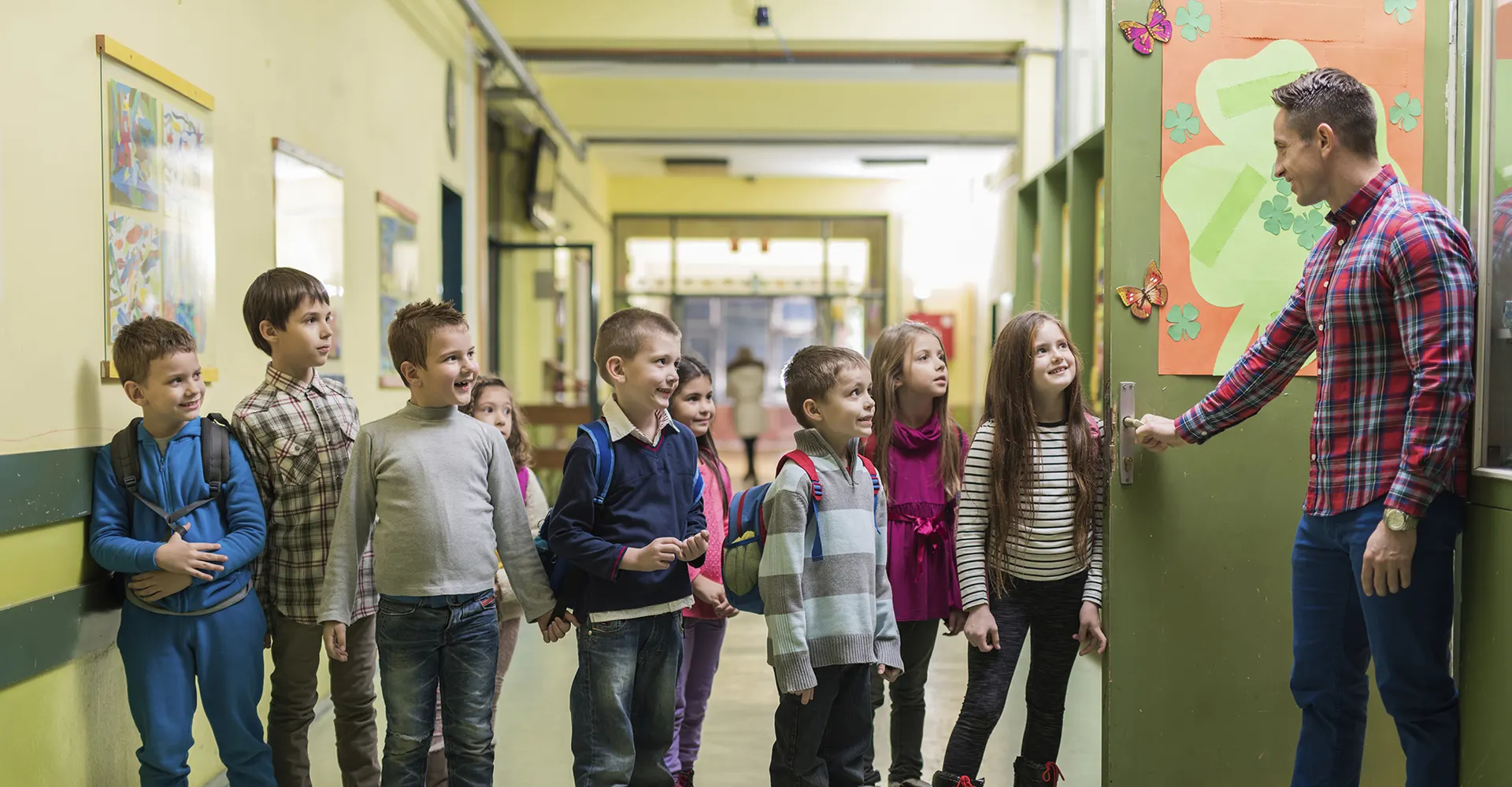 Docente recibiendo en la puerta del salón a una fila de estudiantes de primaria